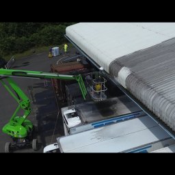 Malowanie dachów, Artdachmal - Pracownik w żółtym kombinezonie na podnośniku czyści zabrudzony dach hali przemysłowej myjką ciśnieniową. Część dachu jest już czysta, widoczny kontrast.