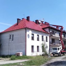 Malowanie dachów, Artdachmal - Budynek z czerwonym dachem w trakcie prac elewacyjnych. Widoczny podnośnik koszowy, drabina oparta o ścianę i świeżo pomalowany dach. Prace wykonywane w słoneczny dzień.
