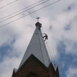 Dwóch alpinistów na szczycie kościoła z cegły, jeden przy krzyżu, drugi na dachu z blachy, liny, błękitne niebo z chmurami w tle, kable elektryczne.