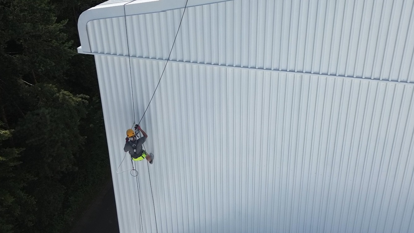 Pracownik na linach, w kasku, przy białej, blaszanej elewacji budynku. Widok z góry, obok drzewa. Praca na wysokościach, zabezpieczenia linowe, jasne oświetlenie.