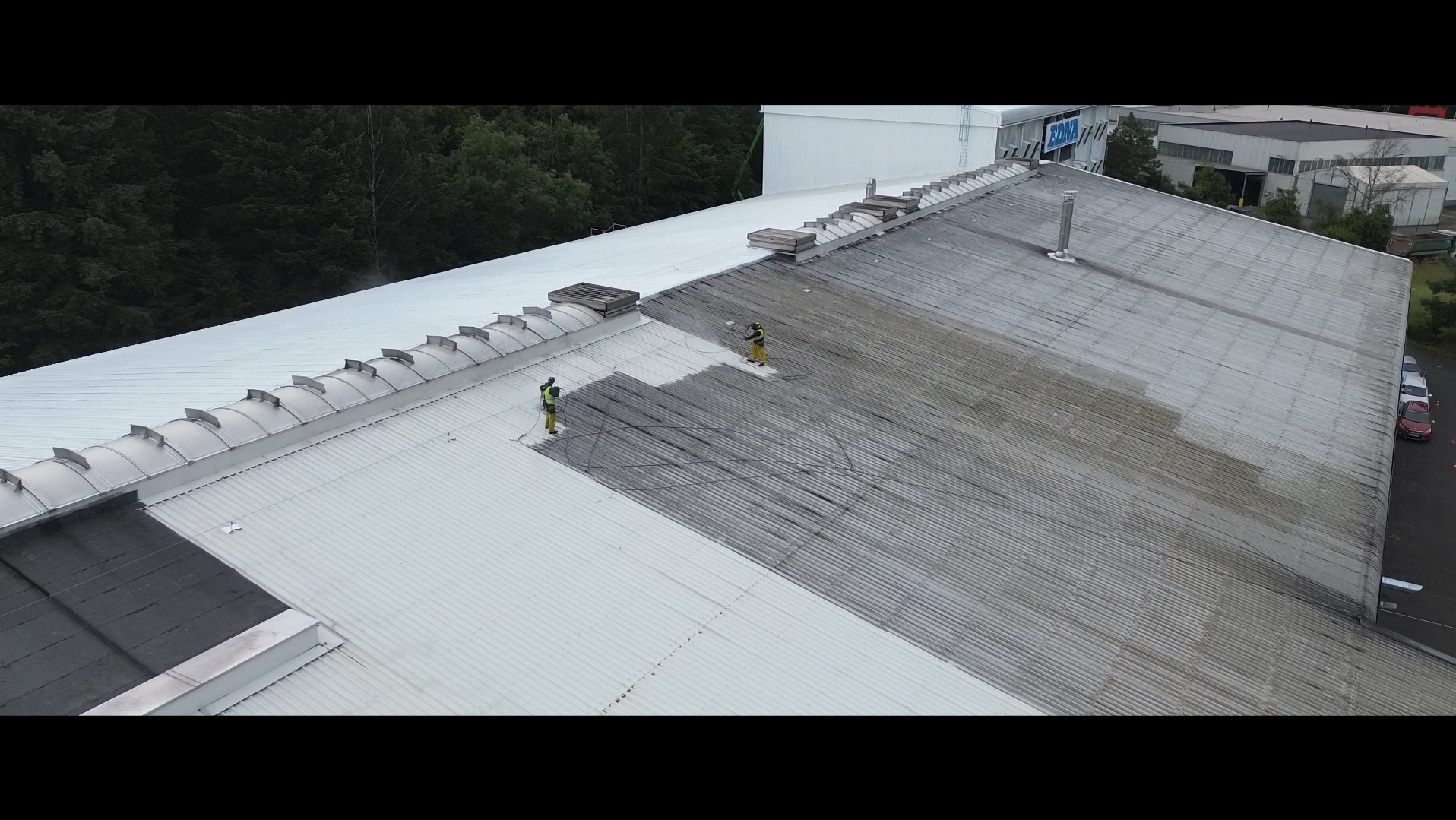 Dwóch pracowników w kombinezonach czyści dach hali przemysłowej za pomocą myjek ciśnieniowych. Widoczny fragment dachu przed i po czyszczeniu.