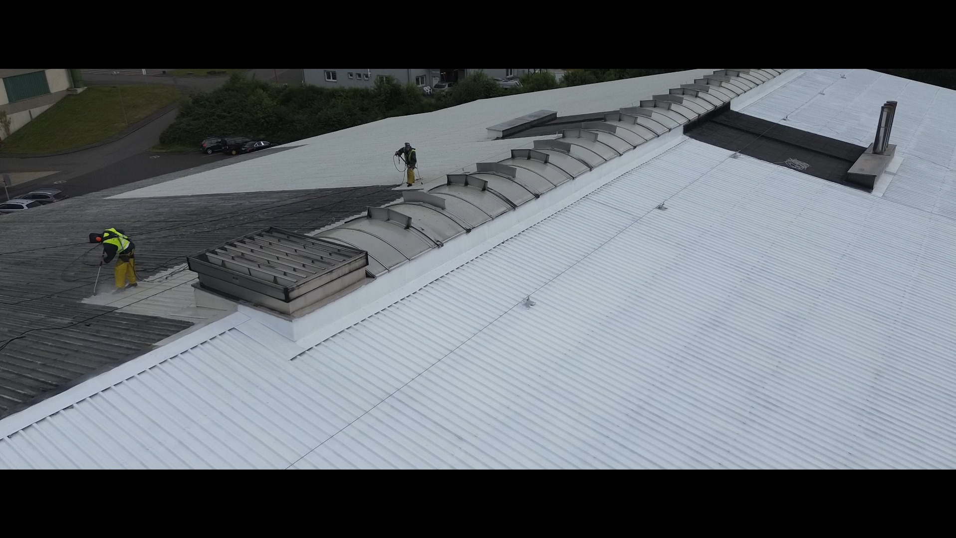 Malowanie dachu przemysłowego z widocznymi świetlikami. Dwóch pracowników w kombinezonach maluje dach na biało, widoczny kontrast między pomalowaną a niepomalowaną częścią dachu.
