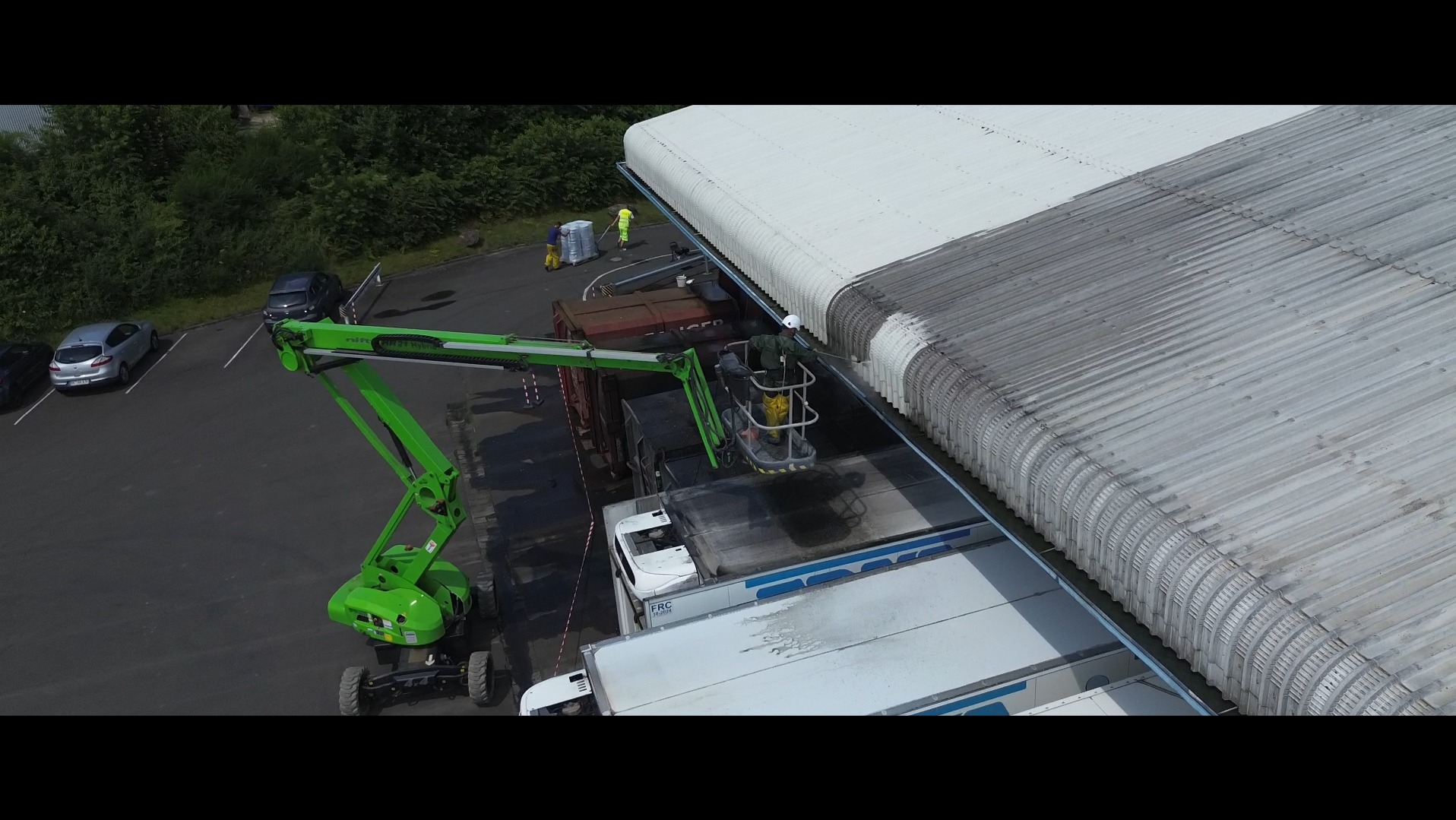 Pracownik w żółtym kombinezonie na podnośniku czyści zabrudzony dach hali przemysłowej myjką ciśnieniową. Część dachu jest już czysta, widoczny kontrast.