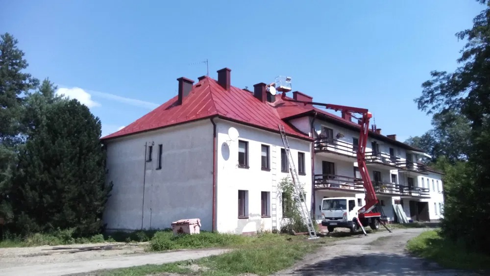 Budynek z czerwonym dachem w trakcie prac elewacyjnych. Widoczny podnośnik koszowy, drabina oparta o ścianę i świeżo pomalowany dach. Prace wykonywane w słoneczny dzień.