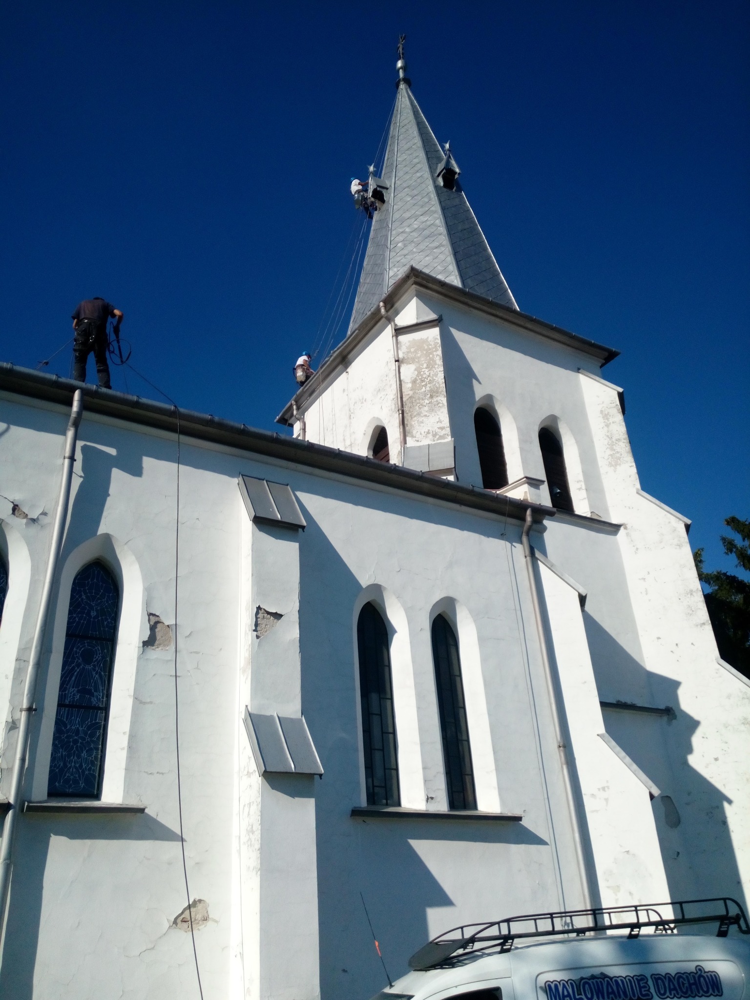 Praca na wysokości: ekipa zabezpieczona linami, remontuje białą elewację kościoła z szarą, metalową wieżą. Na dole kadru widoczny fragment auta z napisem.