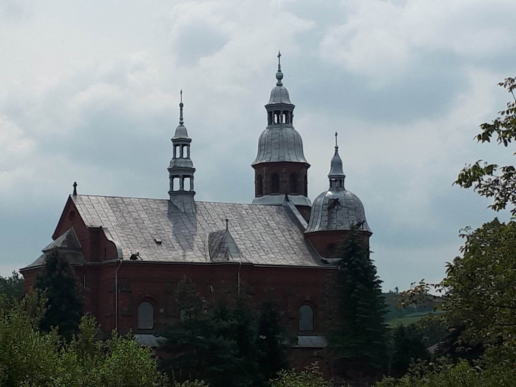 Ceglany kościół z szarym dachem i trzema wieżami na tle pochmurnego nieba, otoczony zielenią drzew. Widoczne uszkodzenia dachu.