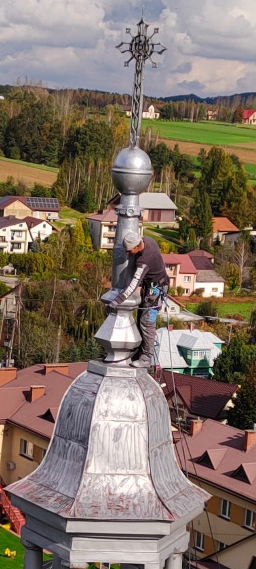 Pracownik zabezpieczony linami, wykonuje obróbki blacharskie na wieży kościoła. W tle zabudowa wiejska i zalesione wzgórza.