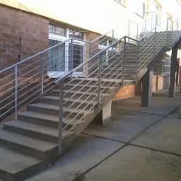 Zewnętrzne schody betonowe z metalową balustradą prowadzące do wejścia na piętrze budynku. Widoczne okna budynku z cegły oraz fragment budynku w kolorze żółtym.