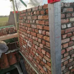 Wznoszenie ściany z czerwonej cegły na budowie domu, widoczne szalunki i pionujące narzędzia z poziomnicą.