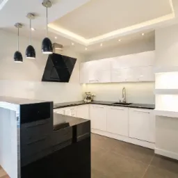 Nowoczesna kuchnia z białymi, lakierowanymi szafkami, czarnym blatem i wyspą z czarnego granitu, oświetlona wiszącymi lampami i podświetleniem LED.