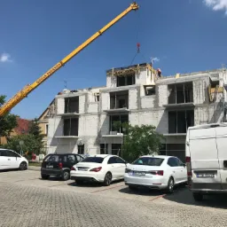Wznoszenie trzypiętrowego budynku z pustaków, z użyciem żółtego dźwigu GRUAS PEDRERA, na tle błękitnego nieba. Na pierwszym planie zaparkowane samochody osobowe i dostawczy.