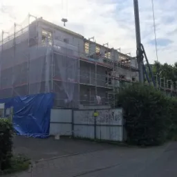 Wznoszenie dwukondygnacyjnego budynku z bloczków, zabezpieczonego siatką i rusztowaniami, widok z ulicy w pochmurny dzień.