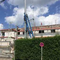 Wznoszenie ścian budynku z białych bloczków, widoczne rusztowania, szalunki na stropie, dźwig w tle, znak drogowy i gęsty żywopłot z przodu.