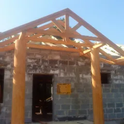 Budowa domu w trakcie: surowa więźba dachowa z jasnego drewna na ścianach z szarego betonu komórkowego, widoczne otwory okienne i drzwiowe. Tablica informacyjna na ścianie.