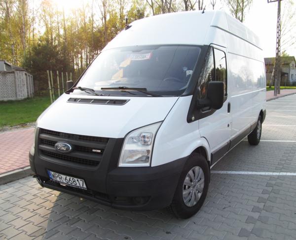 Biały, wysoki bus Ford Transit na parkingu, z widoczną tablicą rejestracyjną i naklejką z imieniem 'Sebastian' na szybie. Słoneczny dzień, drzewa w tle.