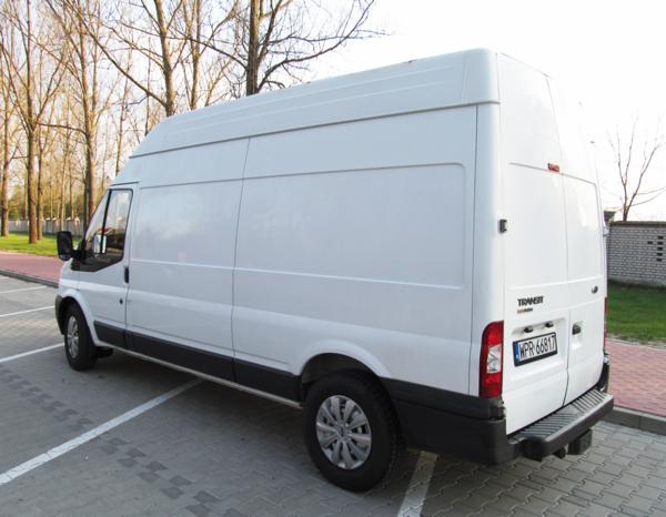 Biały, wysoki bus marki Ford Transit zaparkowany na parkingu z kostki, widok z tyłu i boku. Widoczne drzewa w tle, tablica rejestracyjna.