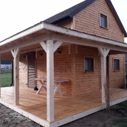 Nowoczesna, drewniana altana z zadaszeniem, połączona z budynkiem mieszkalnym, z drewnianym stołem na tarasie i widokiem na ogród.