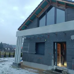 Dom w trakcie ocieplania elewacji styropianem grafitowym, widoczne mocowania, okna z czarnymi ramami, zadaszenie nad wejściem i fragment ogrodu pokrytego śniegiem.