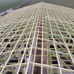 Konstrukcja drewniana dachu domu w trakcie budowy, widok z góry na siatkę belek z metalowymi łącznikami, na tle zielonego pola i ogrodzenia.