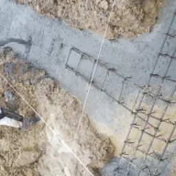 Wylewanie betonu do wykopu z ułożonym zbrojeniem, widoczne fragmenty ubrania robotnika i wąż do betonu.