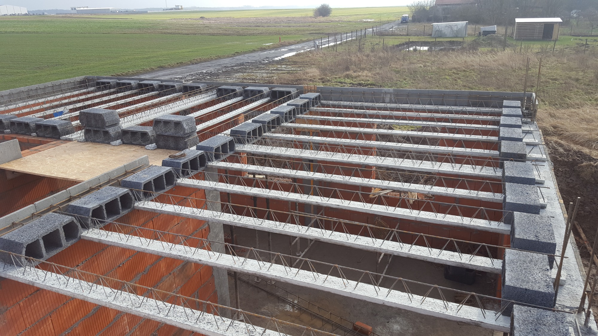 Wieniec stropowy z betonowych bloczków i kratownic stalowych na murach z czerwonej cegły w trakcie budowy domu jednorodzinnego, widok z góry na tle krajobrazu polnego.