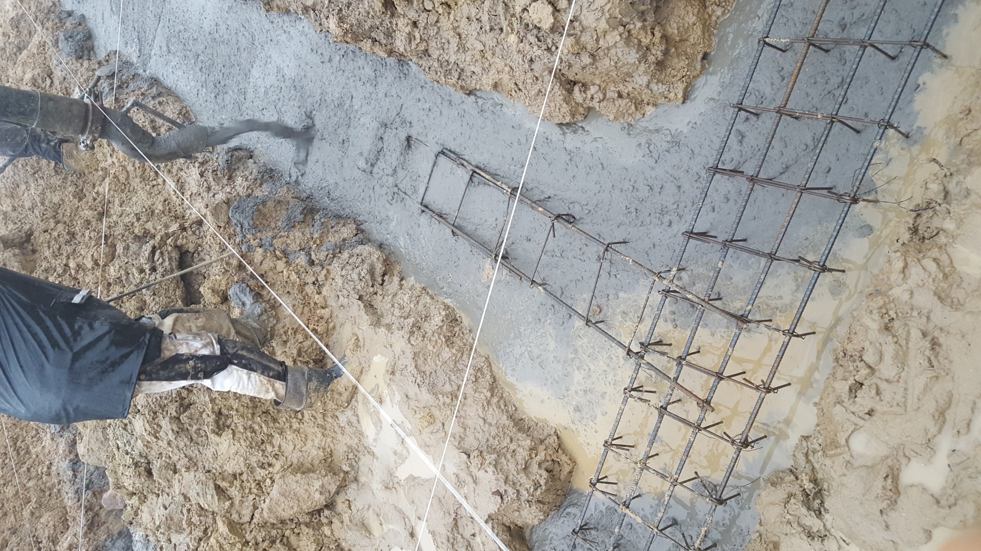 Wylewanie betonu do wykopu z ułożonym zbrojeniem, widoczne fragmenty ubrania robotnika i wąż do betonu.