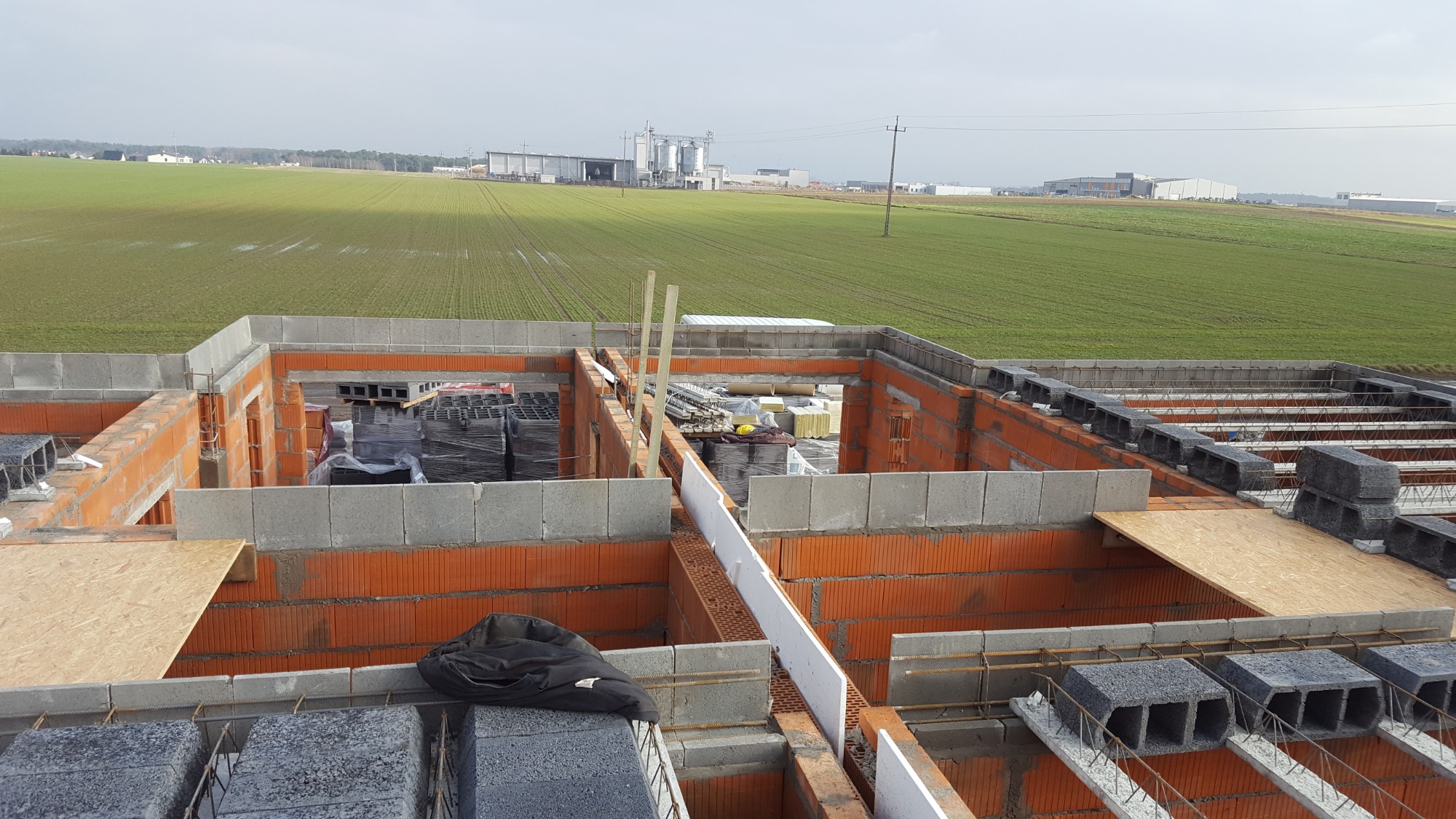 Wznoszenie ścian parteru domu z betonu komórkowego i cegły ceramicznej, widok z góry na budowę w otoczeniu pól uprawnych i zabudowań gospodarczych.