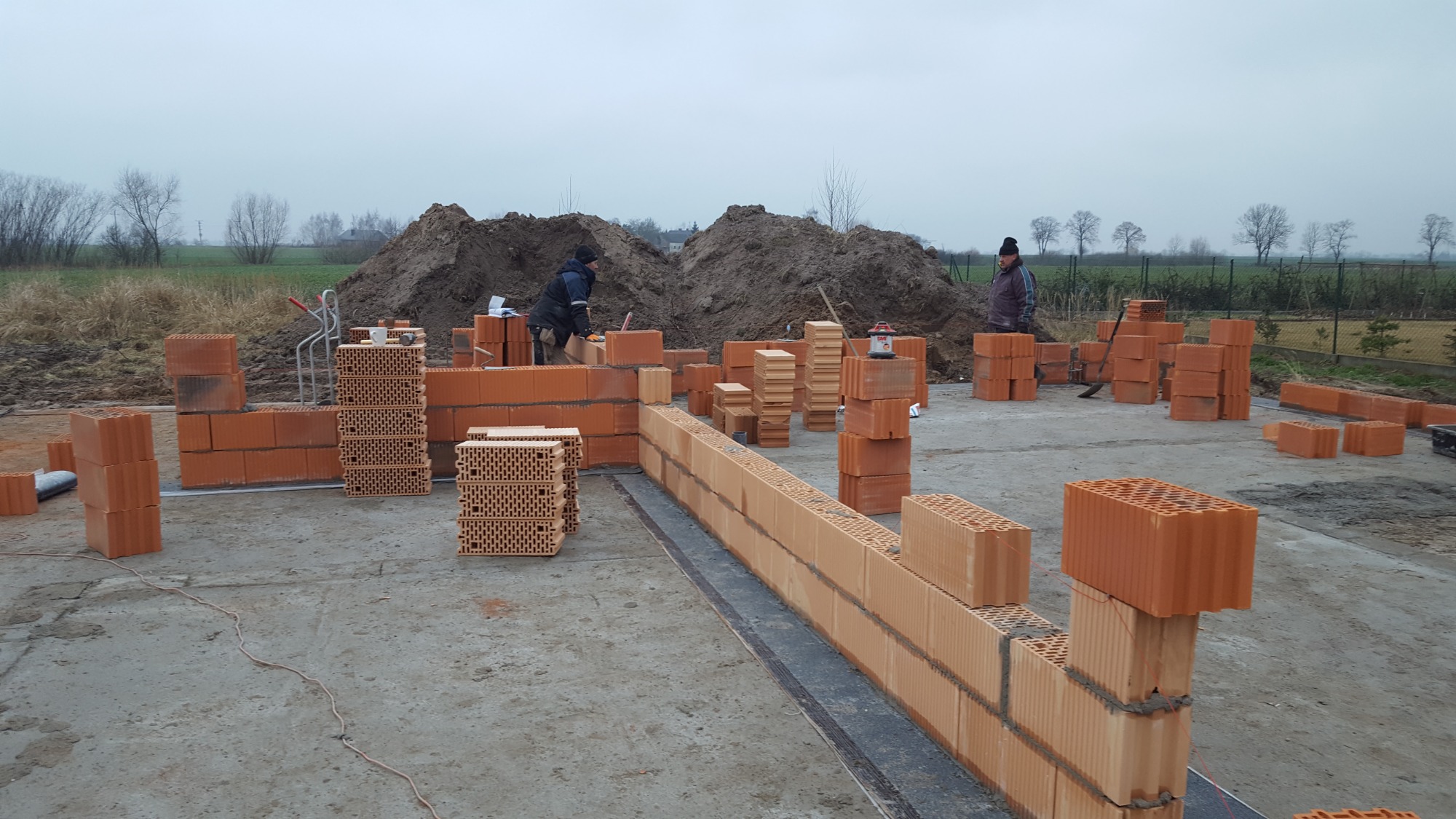 Wznoszenie muru z ceramicznych pustaków na fundamencie, widoczni dwaj pracownicy w trakcie pracy, na drugim planie hałda ziemi i drzewa na tle pochmurnego nieba.