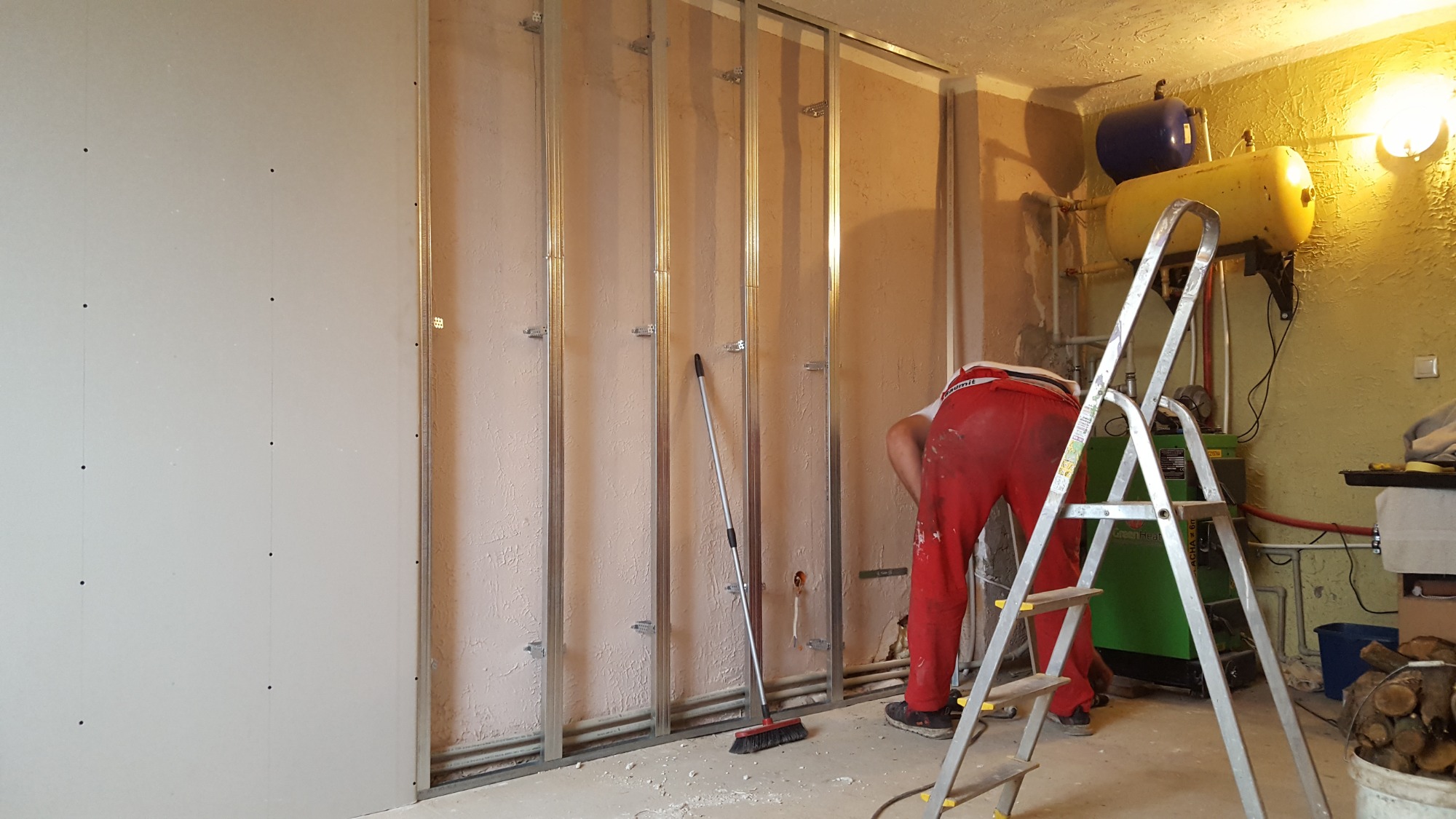 Montaż płyty karton gips na metalowym stelażu w pomieszczeniu z instalacją grzewczą, widoczna drabina i pracownik w czerwonych spodniach.