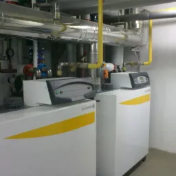 Dwa białe kotły grzewcze De Dietrich z żółtymi akcentami, widoczne rury i zawory instalacji. Ujęcie w kotłowni, z bliska, pokazujące detale instalacji.