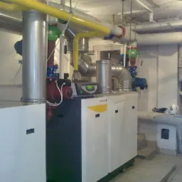 Złożona instalacja grzewcza z kotłem De Dietrich, rurami, pompami i zaworami w kotłowni. Widoczne elementy instalacji w kolorze żółtym, czerwonym i srebrnym.