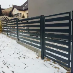 Nowoczesne ogrodzenie z poziomych, szarych paneli na betonowej podmurówce, częściowo pokryte śniegiem, z widocznym fragmentem domu w tle.
