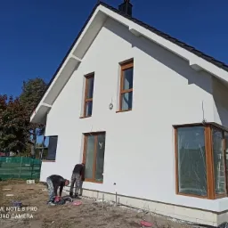 Dwóch pracowników przygotowuje elewację domu jednorodzinnego z drewnianą stolarką okienną, widoczne narzędzia i materiały budowlane.