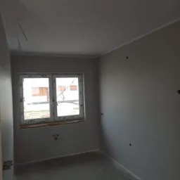 Surowe wnętrze pokoju po nałożeniu pierwszej warstwy szarej gładzi, widoczne przygotowane punkty elektryczne, okno PCV i wystające przewody.