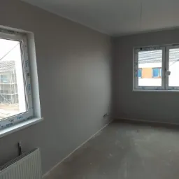 Pomieszczenie po malowaniu ścian na szaro, widoczne dwa okna z białymi ramami marki VEKA, grzejnik i surowa podłoga.
