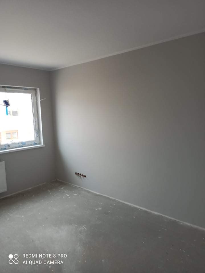 Pomalowane na szaro wnętrze pokoju z widocznymi gniazdkami elektrycznymi, surową podłogą i oknem z widokiem na zewnątrz.