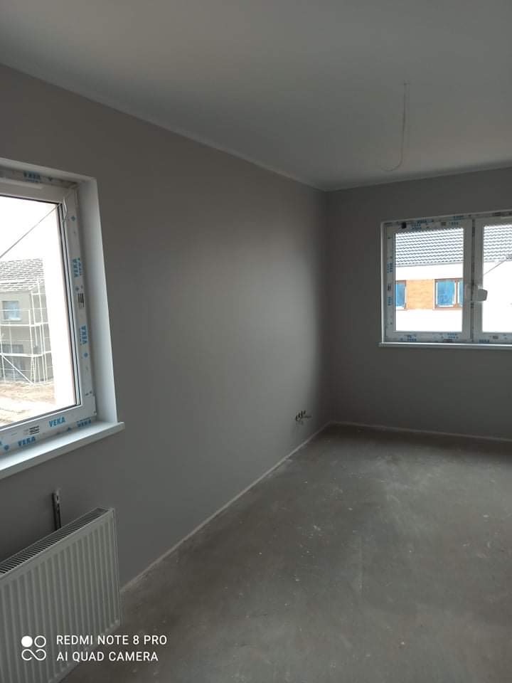 Pomieszczenie po malowaniu ścian na szaro, widoczne dwa okna z białymi ramami marki VEKA, grzejnik i surowa podłoga.