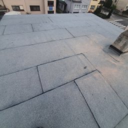 ART-BUD - Płaski dach pokryty papą termozgrzewalną z widocznym kominem i fragmentem buta w rogu kadru, w tle zabudowa miejska.