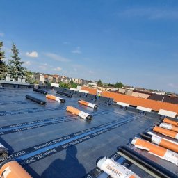 ART-BUD - Rozwinięte rolki papy termozgrzewalnej BauderTEC Sprint DUO na płaskim dachu w trakcie prac dekarskich, widok z góry na tle zabudowy miejskiej i błękitnego nieba.