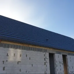 Nowy, grafitowy dach z dachówki ceramicznej na surowym budynku z białego bloczka, widoczna pomarańczowa rura spustowa.