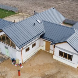 ART-BUD - Dom w trakcie budowy z nowym, szarym dachem z blachy na rąbek stojący, widok z drona, z widocznymi rynnami i oknami z drewnianą stolarką, teren wokół w trakcie przygotowania.