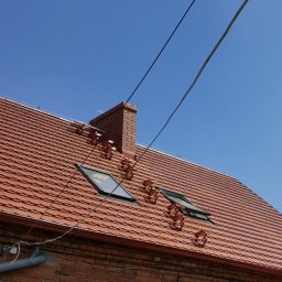 ART-BUD - Czerwony dach z dachówki ceramicznej z dwoma oknami dachowymi, kominem i uchwytami przeciwśniegowymi, widok z dołu na tle błękitnego nieba.