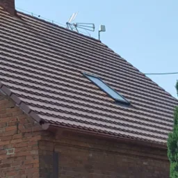 Czerwony dach z dachówki ceramicznej na ceglanym domu z oknem dachowym i anteną telewizyjną na szczycie.