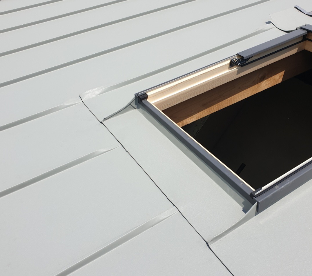 Jasnoszary dach z blachy trapezowej z otworem na okno dachowe, widoczna drewniana konstrukcja i precyzyjne obróbki blacharskie wokół otworu.