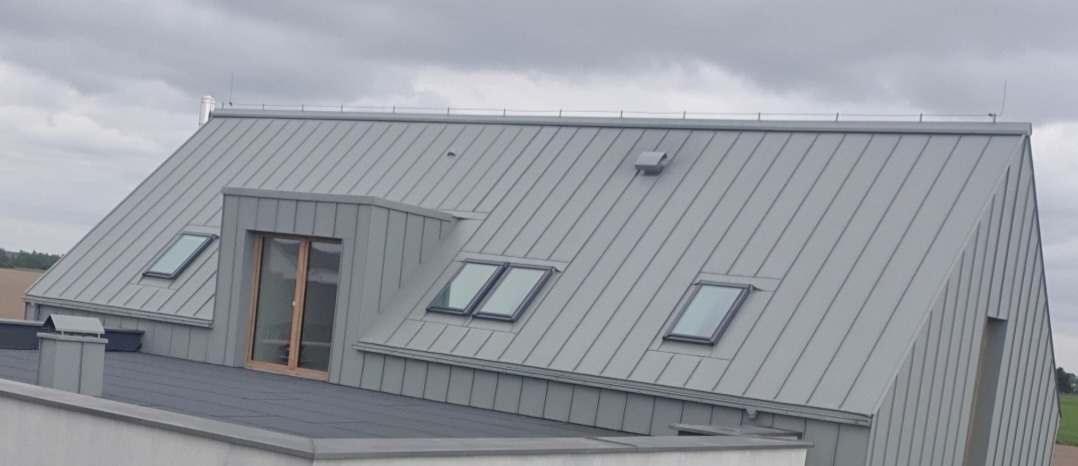 Szary dach z blachy na nowoczesnym domu z oknami dachowymi i lukarną z drzwiami balkonowymi.