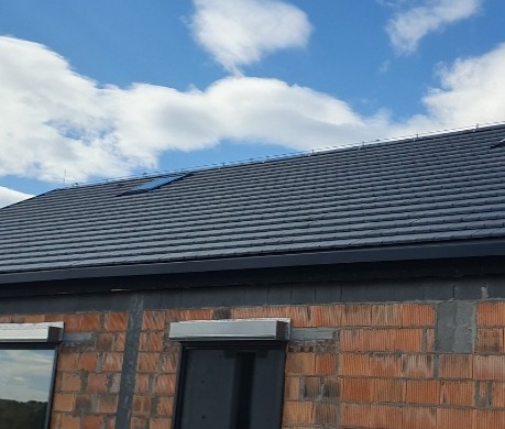 Nowy dach z ciemnej dachówki ceramicznej z oknem połaciowym, widoczny fragment murowanej ściany budynku z oknami.