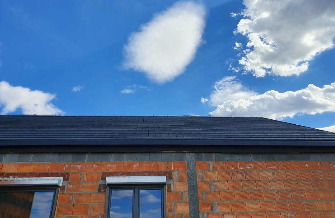 Nowy, ciemnoszary dach z dachówki ceramicznej nad surowym murem z cegły ceramicznej porotherm, błękitne niebo z białymi chmurami odbija się w szybach okien.