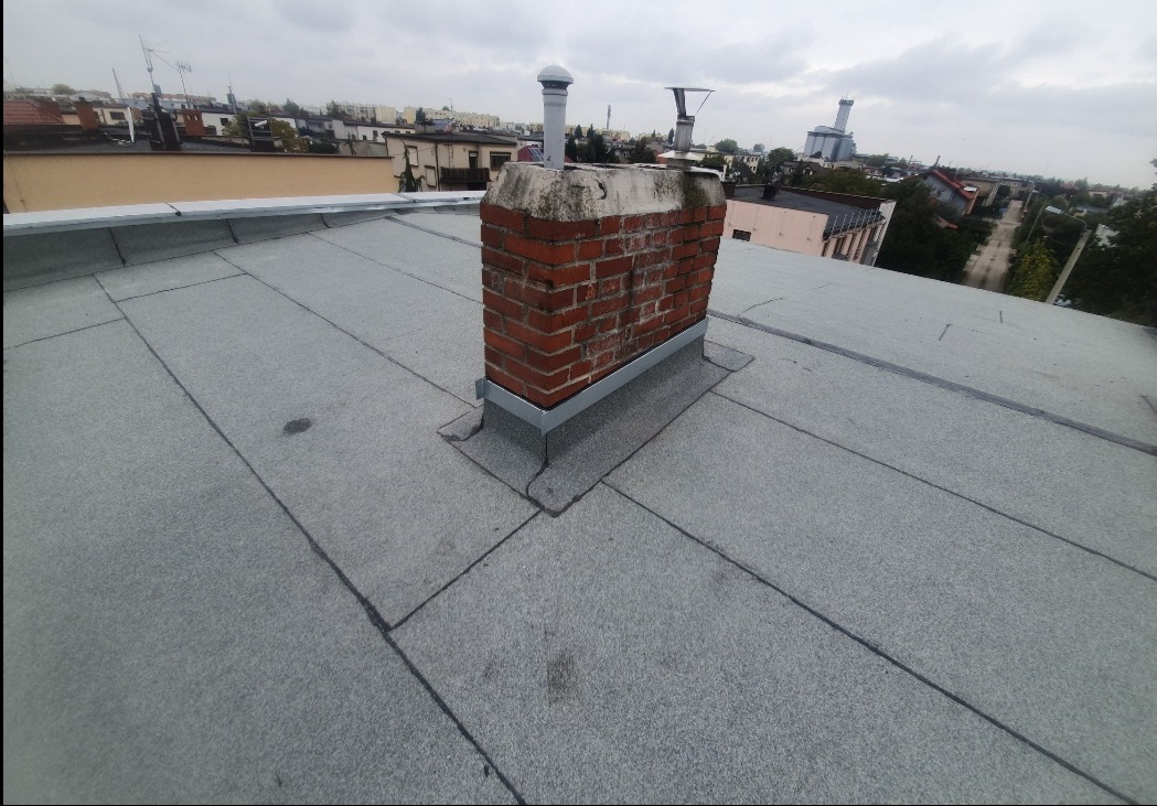 Płaski dach pokryty szarym materiałem bitumicznym, z widocznym kominem obłożonym blachą, na tle zabudowy miejskiej w pochmurny dzień.