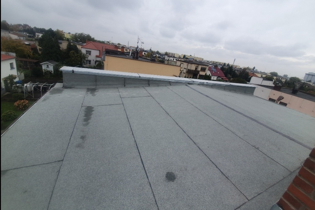 Płaski dach pokryty papą termozgrzewalną, widoczne łączenia i obróbki blacharskie na krawędzi, w tle zabudowa miejska i pochmurne niebo.