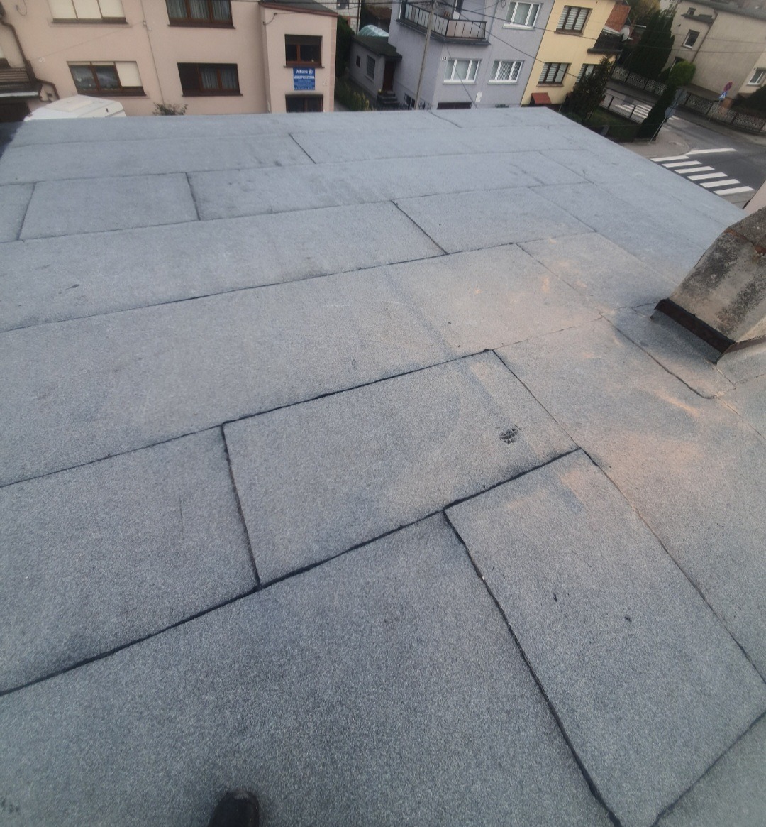 Płaski dach pokryty papą termozgrzewalną z widocznym kominem i fragmentem buta w rogu kadru, w tle zabudowa miejska.